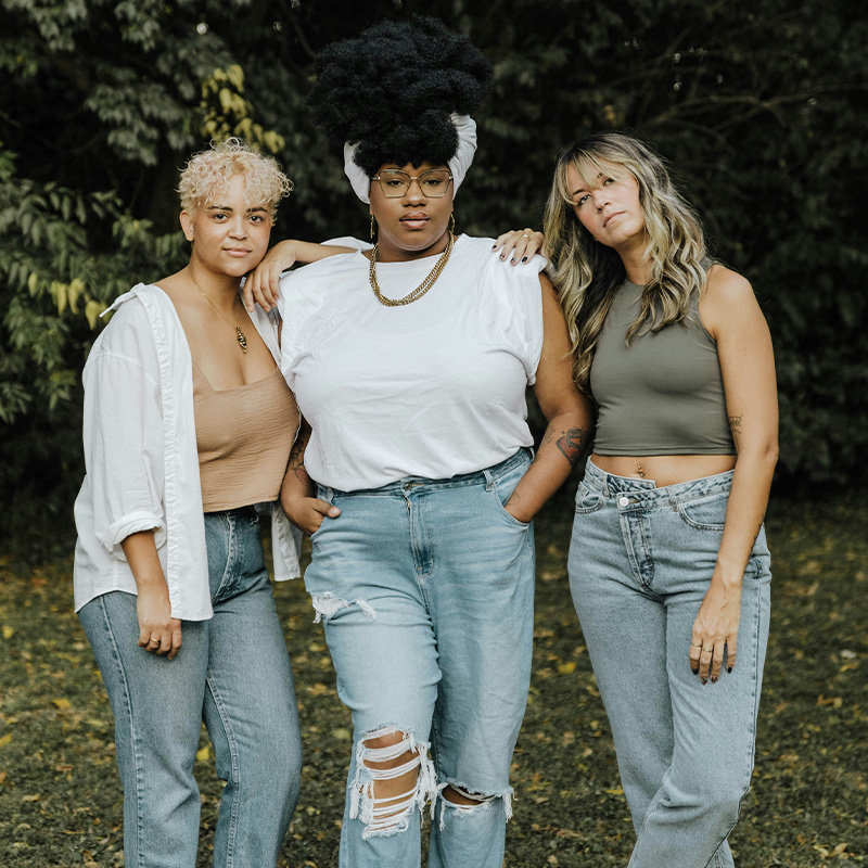 Three women pose together outdoors, dressed in casual outfits, against a green, leafy backdrop.