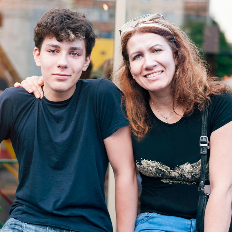 A mother and son sitting next to each other, smiling at the camera, the mother has her hand on his shoulder.