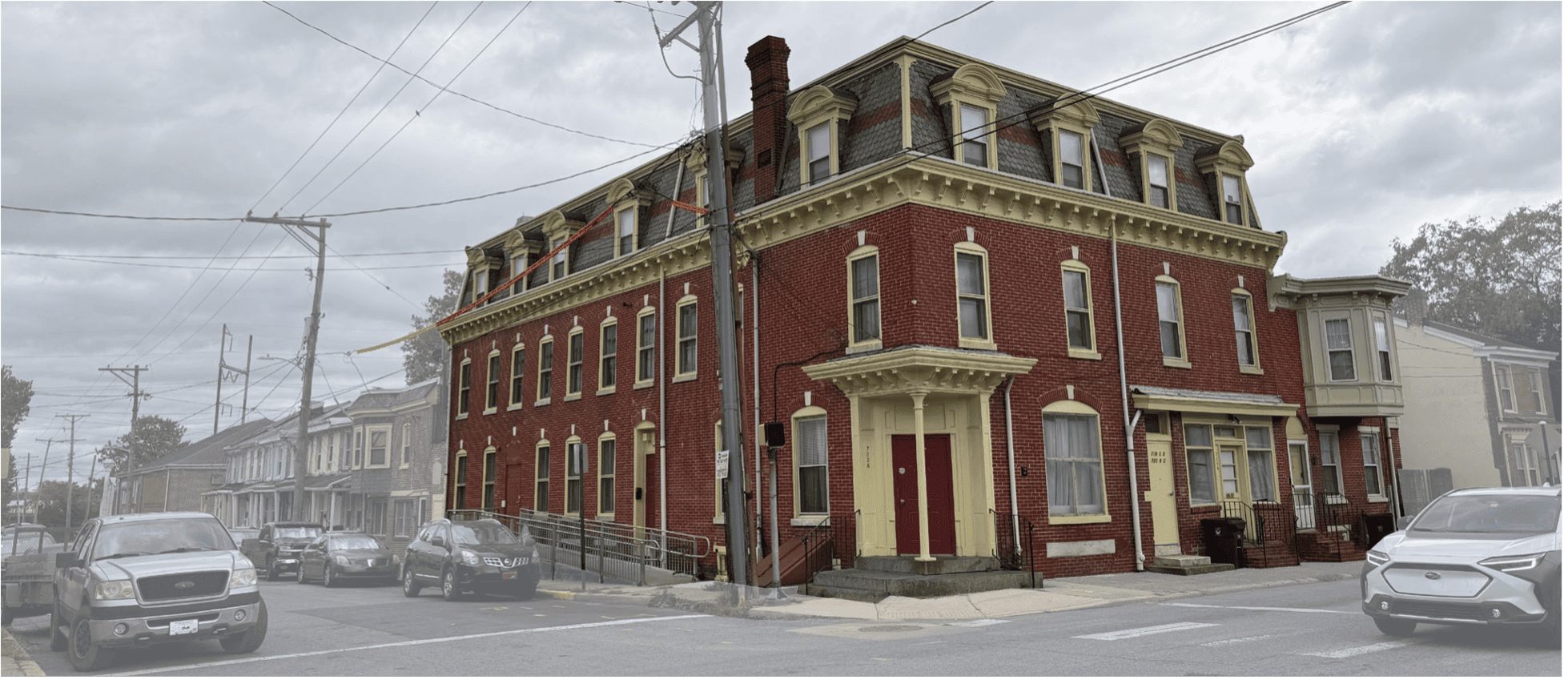 A historic three-story brick building with yellow trim, surrounded by vehicles and power lines under a cloudy sky.