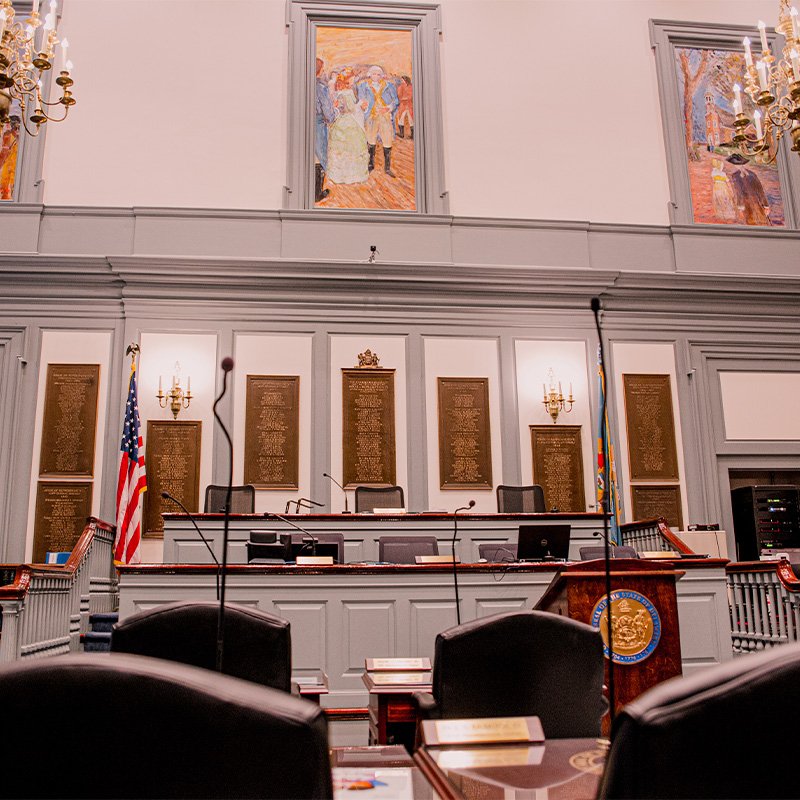 Interior of the Legislative Hall chamber featuring a podium, empty seats, framed artwork, a U.S. flag, and bronze plaques on the wall.
