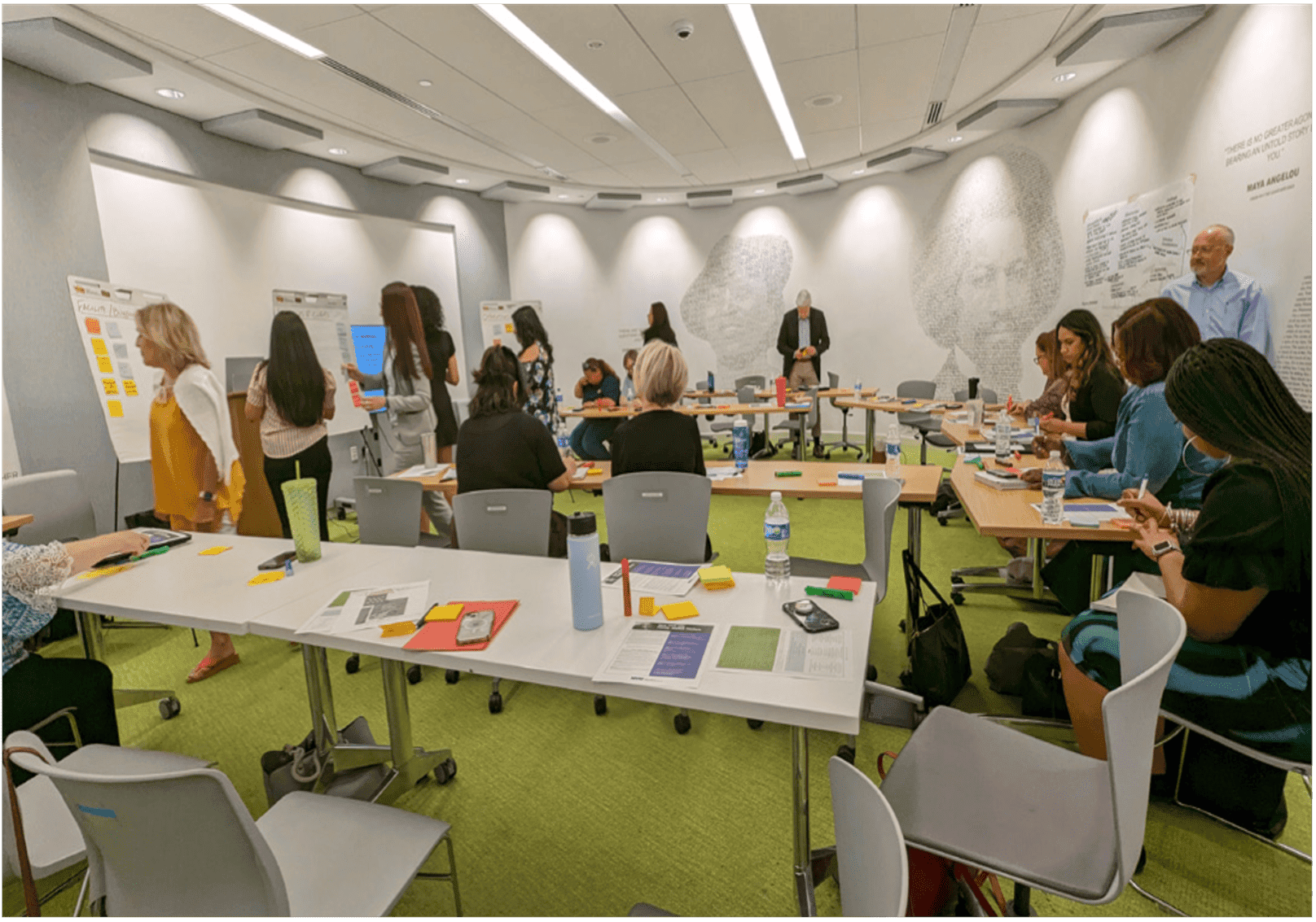 Group of people working and a white room at tables and placing post-it notes on multiple boards.