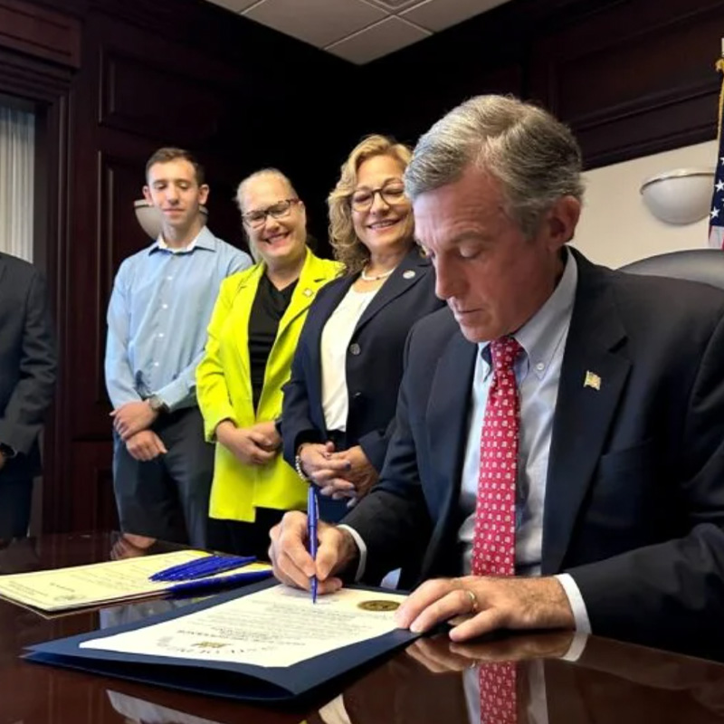Governor John Carney signing legislation with multiple people standing smiling beside him.